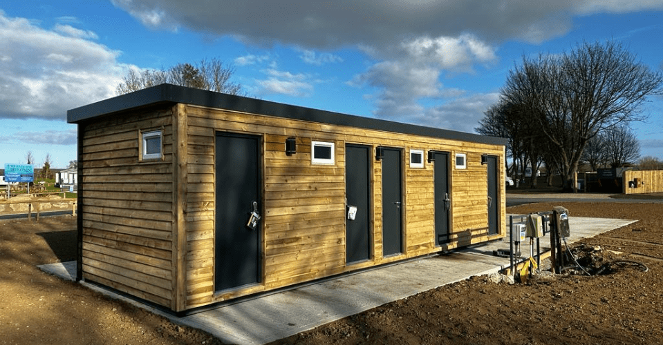 A wooden building with four black doors stands on a concrete pad in an open area with patches of grass and trees under a partly cloudy sky.