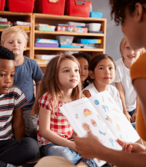A teacher reads a picture book to an attentive group of young children sitting in a classroom with shelves of colorful storage bins in the background.