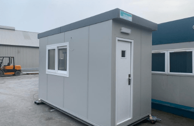 A small, grey modular building with a door and window sits on an industrial lot with other similar structures and a forklift in the background.